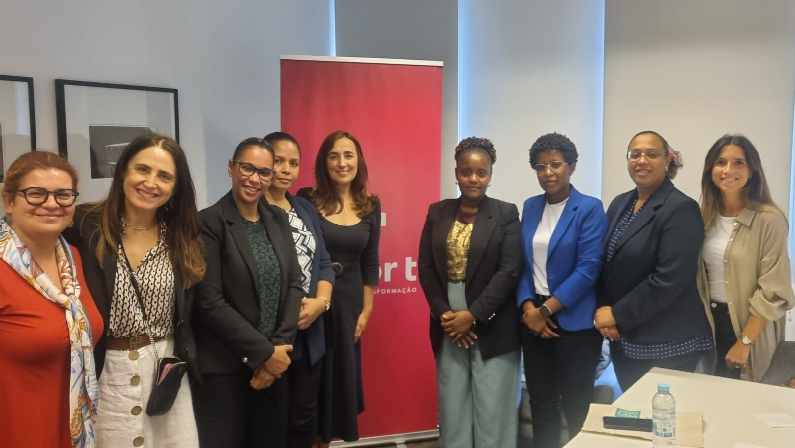 fotografia ao grupo da delegação de cabo verde. Na fotografia, estão 9 mulheres de diferentes idades em fila, viradas para a camara. a fotografia foi tirada numa sala, tendo um roll-up da Valor T atrás das pessoas.